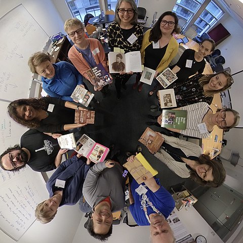 Photograph of Smithsonian staff and volunteers at a Wikipedia edit a thon to increase the representation of women in Wiki records. The group is standing in a circle, looking up at the camera and smiling, and. holding historic photographs of women they recorded information on.