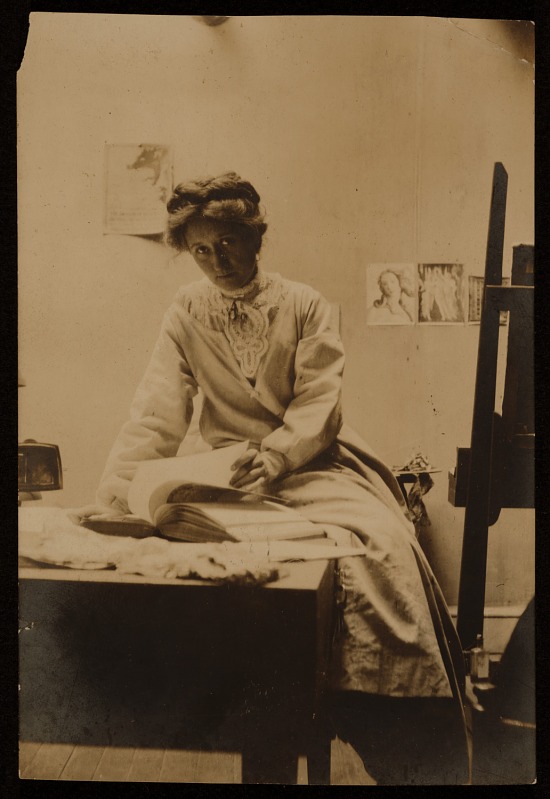 Sepia toned image of artist Olive Rush. She is sitting in an art studio, with an easel in front of her and she is paging through a large book.